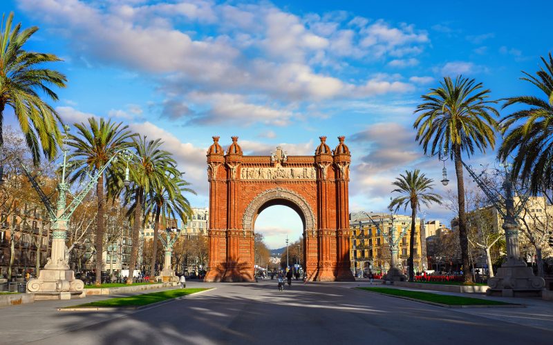 Triumphal Arch in Barcelona, Catalonia, Spain. Arc de Triomf at boulevard street. Alley with tropical palm trees. Early morning landscape with shadows and blue sky with clouds. Famous landmark.