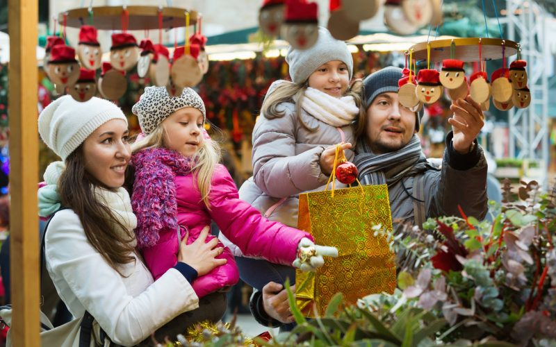 Happy smiling family of four buying Caga Tio at Christmas market. Focus on woman and ittle girl